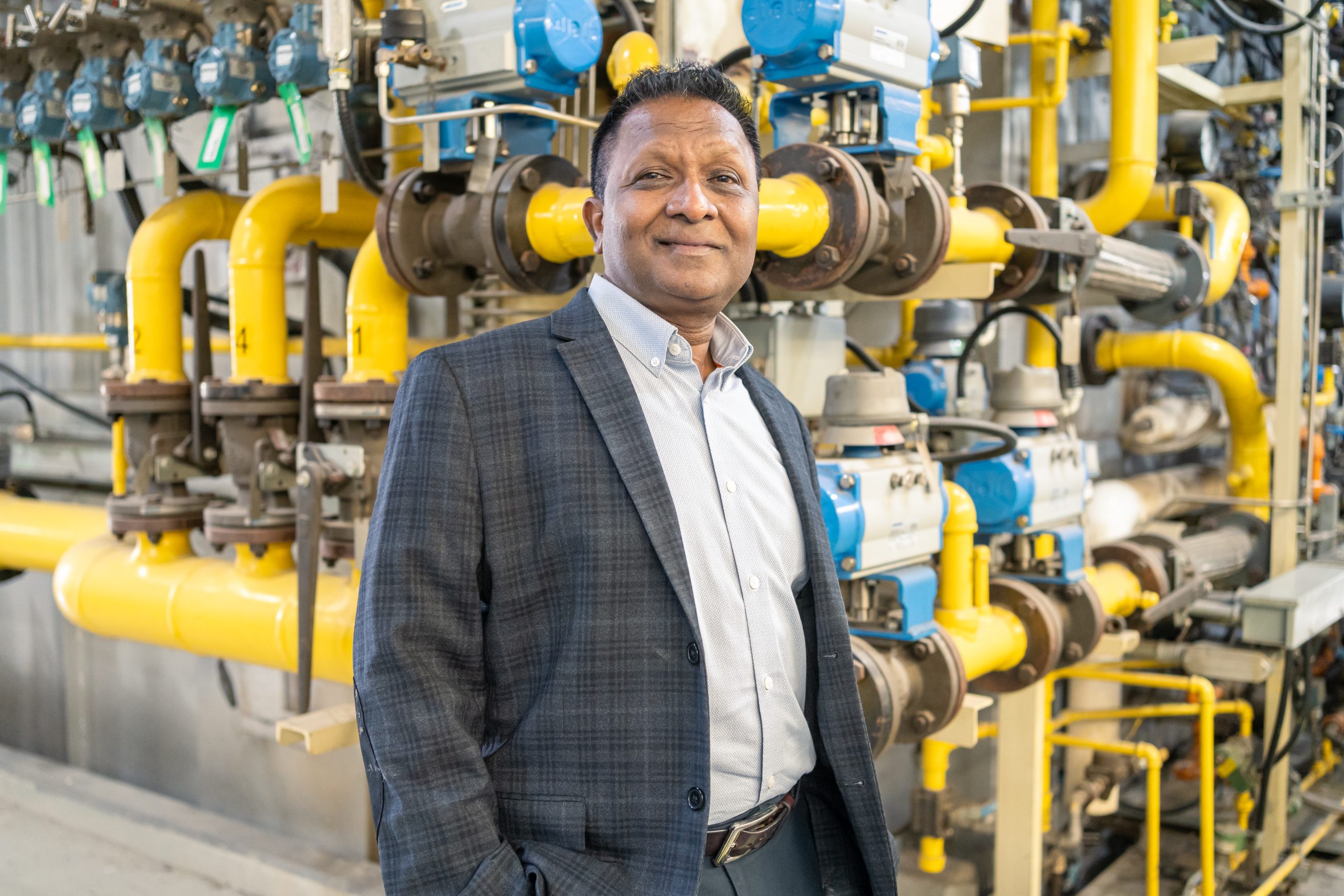 A photo of Rajesh Patel standing in the central steam plant in a jacket and dress shirt, smiling.