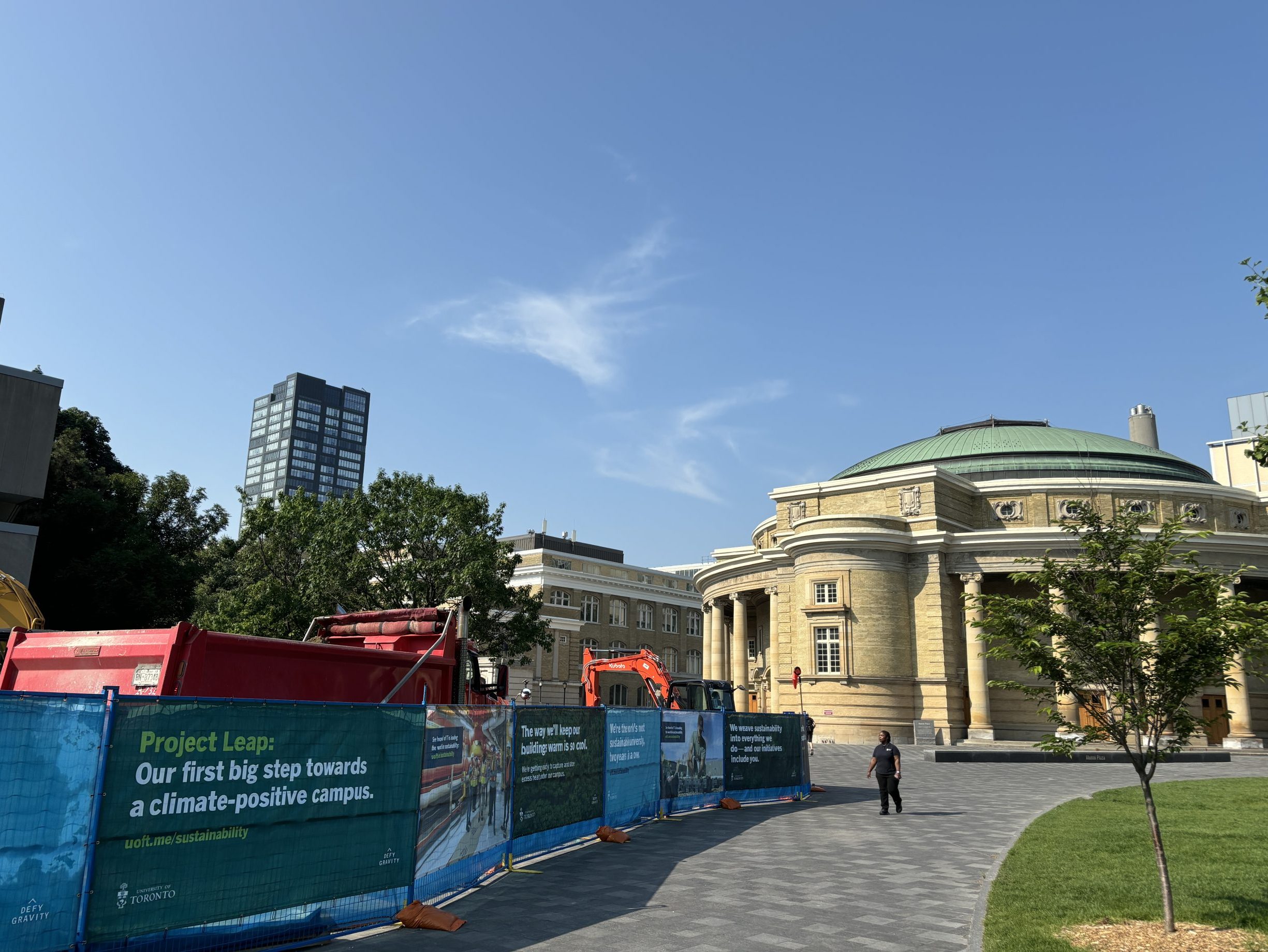 A photo of Kings College Circle, looking toward Convocation Hall. A new tree is on the field, and on the other side of the walkway is a row of display panels with construction vehicles visible behind them. The display panel closest to the viewer is green with text that reads "Project Leap: Our first big step towards a climate-positive campus"