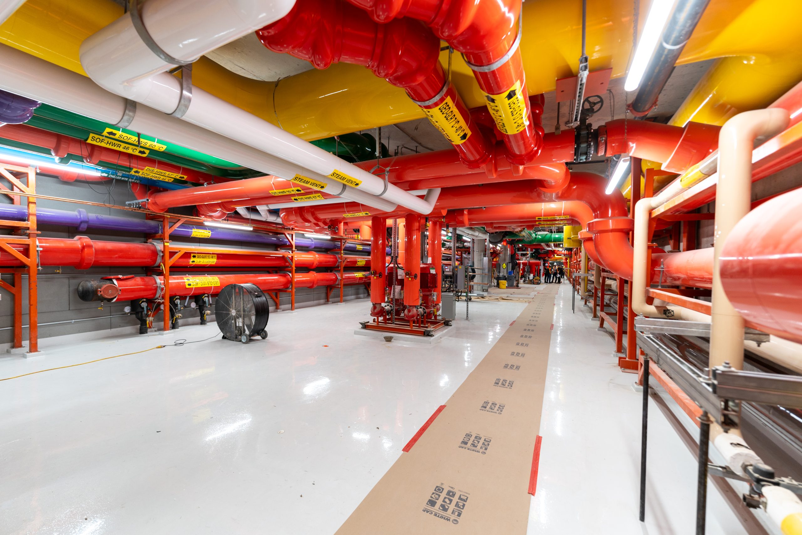 A view inside the brightly lit geoexchange mechanical room, with the walls and ceilings covered in wide, colourful pipes. The floor is white with a cardboard walkway set up. 