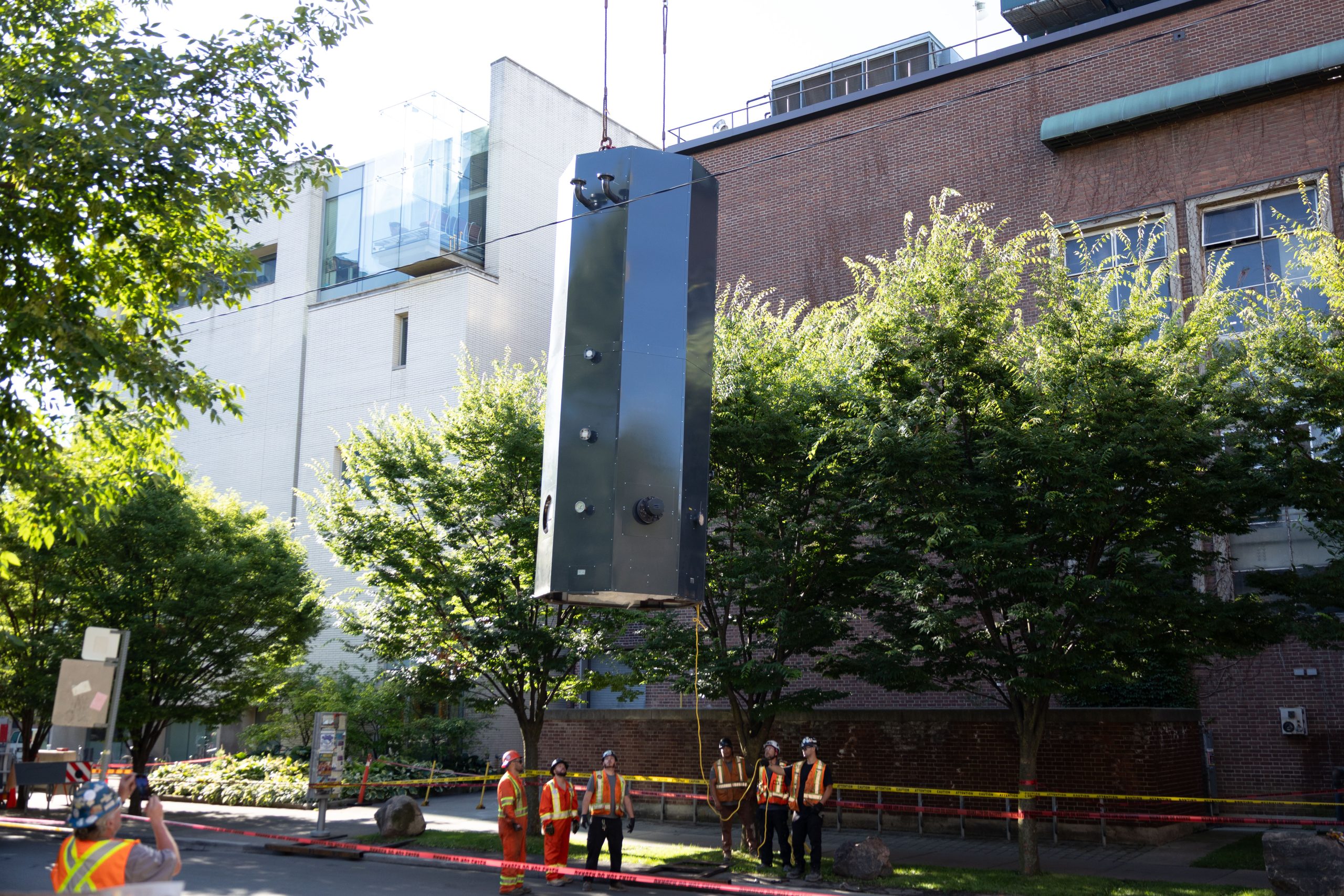 A photo of the boiler, about the height of four people, being lifted in the air. The crane is not visible in the shot, but the side of the central steam plant is, as is a group of people in construction protective equipment standing below the boiler as it rises into the air.