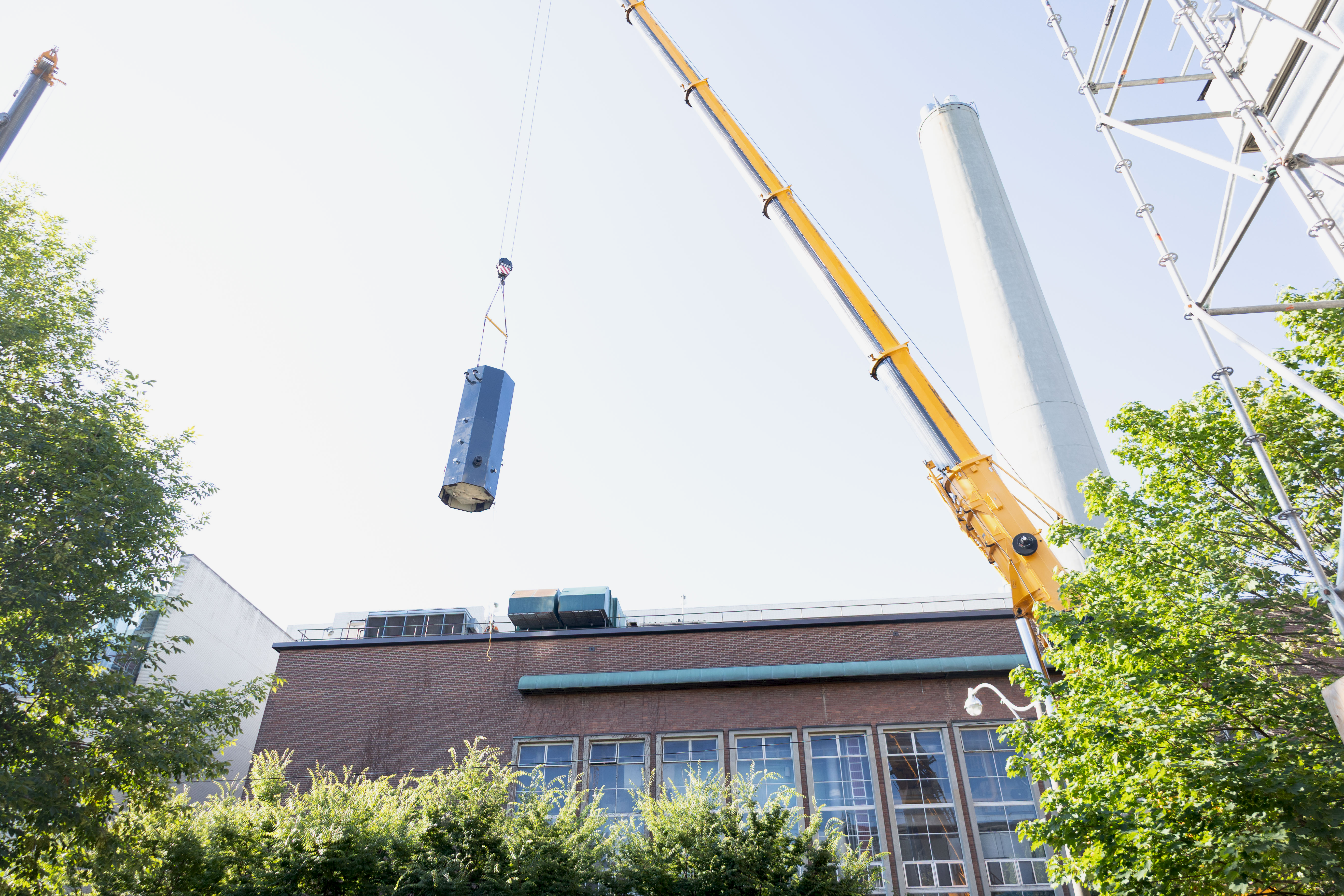 A photo of the outside of the central steam plant, looking up. A crane is lifting a large dark metal cylinder into the air, above the room of the steam plant. The smoke stack and the trees surrounding the plant are also visible.