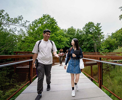 Two students walk on a raised pathway, surrounded by greenery, smiling and engaged in conversation.